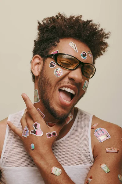 Expressive african american fella in sunglasses with trendy stickers on face on grey background — Stock Photo
