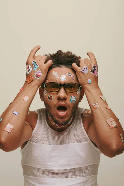 Shocked young african american man in sunglasses with trendy stickers on face on grey background — Stock Photo