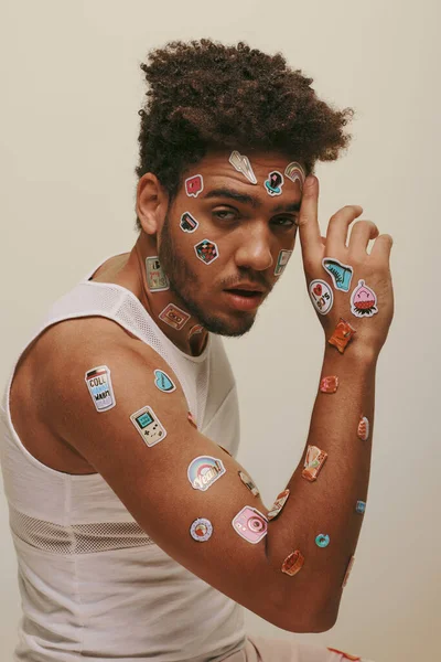 Cool homme afro-américain en débardeur avec des autocollants sur le visage en regardant la caméra sur fond gris — Photo de stock