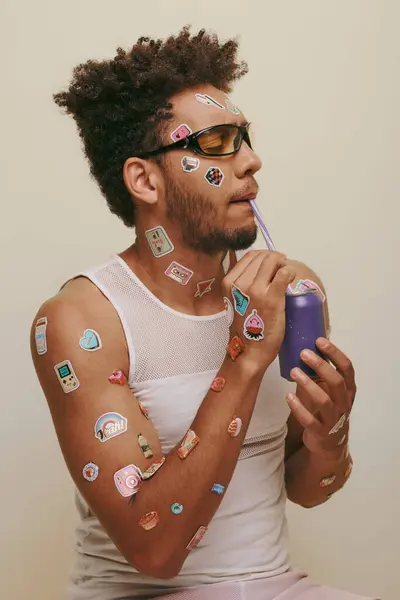 Hombre afroamericano rizado con pegatinas en la cara y el cuerpo beber soda sobre fondo gris - foto de stock