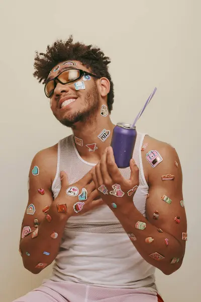 Cheerful african american man with stickers on face and body holding soda can on grey background — Stock Photo