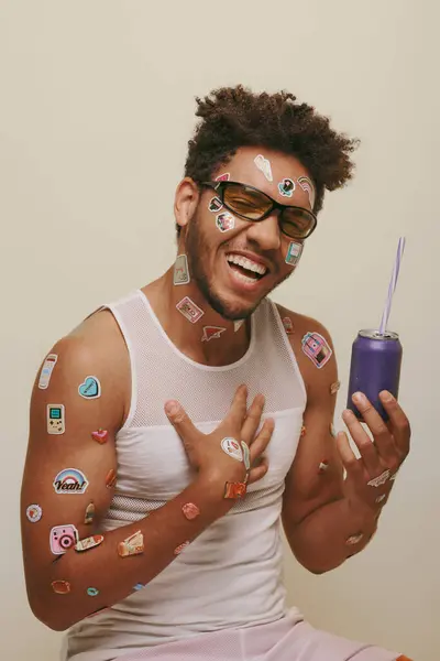 Hombre afroamericano feliz con pegatinas en la cara y el cuerpo sosteniendo lata de refresco sobre fondo gris - foto de stock