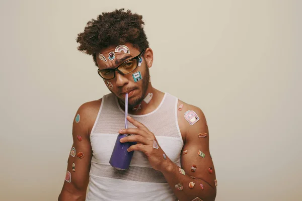 Joven afroamericano chico con pegatinas en la cara y cuerpo beber soda sobre fondo gris - foto de stock