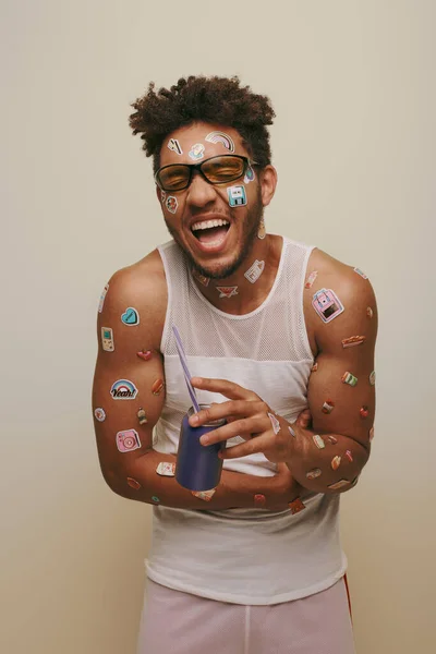 Hombre afroamericano excitado con pegatinas en la cara y el cuerpo sosteniendo lata de refresco sobre fondo gris - foto de stock