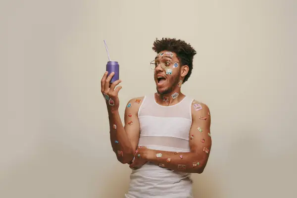 Homme afro-américain étonné avec des autocollants sur le visage et le corps en regardant la canette de soda sur fond gris — Photo de stock