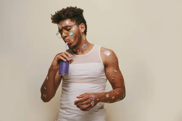 Joven y rizado afroamericano chico con pegatinas en la cara y el cuerpo beber soda sobre fondo gris - foto de stock