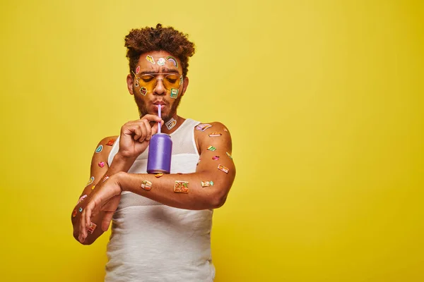 Curly african american guy with stickers on face and body drinking soda on yellow background — Stock Photo