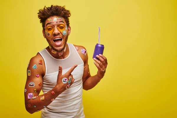 Hombre afroamericano excitado con pegatinas en la cara apuntando a lata de refresco sobre fondo amarillo - foto de stock