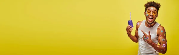 Збуджений афроамериканський чоловік з наклейками на обличчі, що вказують на содову банку на жовтому фоні — стокове фото