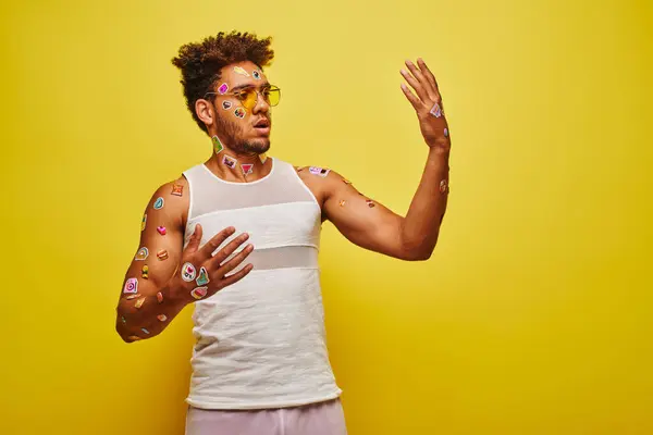 Hombre afroamericano sorprendido mirando pegatinas en sus manos y cuerpo sobre fondo amarillo - foto de stock