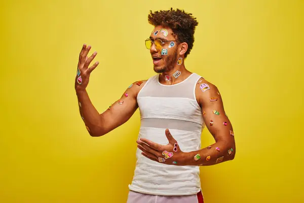 Asombrado hombre afroamericano mirando pegatinas en sus manos y cuerpo sobre fondo amarillo - foto de stock