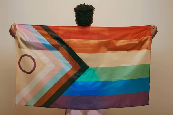 Vista posterior del hombre afroamericano sosteniendo la bandera del arco iris lgbt sobre fondo amarillo, mes del orgullo - foto de stock