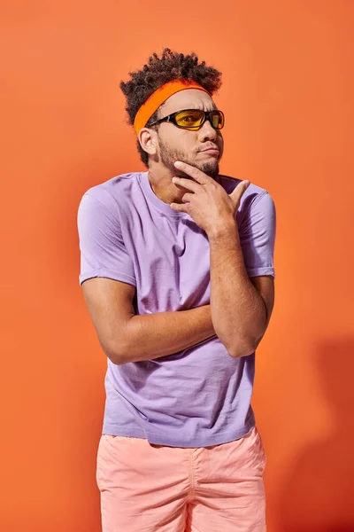 Réfléchi jeune homme afro-américain en bandeau et lunettes de soleil sur fond orange, drôle de visage — Photo de stock