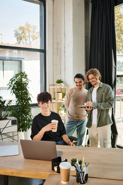 Alegre joven con café para ir usando el ordenador portátil cerca de compañeros de trabajo masculinos discutiendo proyecto de inicio - foto de stock