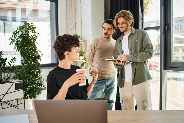 Young man with coffee to go pointing at laptop and looking at coworkers while discussing project — Stock Photo