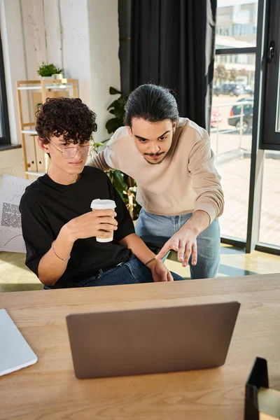 Joven apuntando a la computadora portátil mientras explica su idea de proyecto a su compañero de trabajo con café para ir - foto de stock