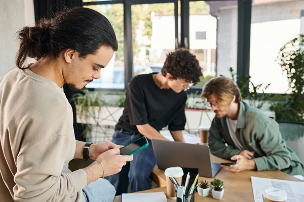 Young professional using smartphone near blurred colleagues brainstorming over a laptop in office — Stock Photo