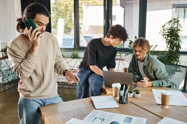 Young man having phone call and looking at charts near blurred coworkers brainstorming over laptop — Stock Photo