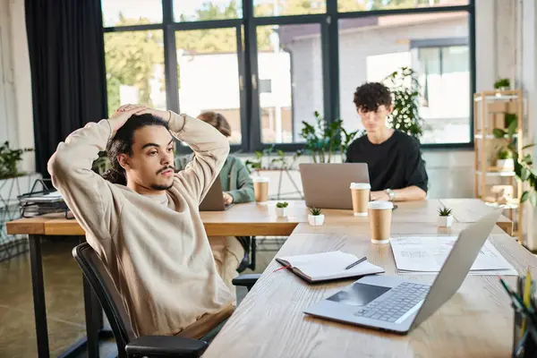 Jeune entrepreneur fatigué analyser les données sur ordinateur portable avec des collègues flous dans un espace de travail moderne — Photo de stock