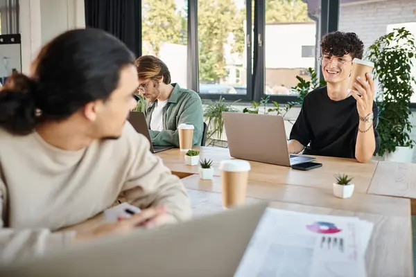 Happy young men working on laptops in a coworking space, man in 20s offering coffee to colleague — Stock Photo