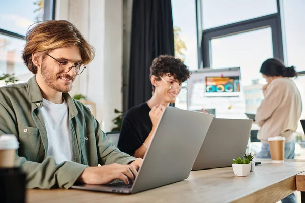 Cheerful young startup team smiling while working on laptops in modern coworking space, men in 20s — Stock Photo