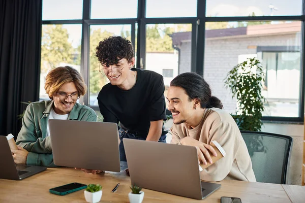 Smiling young men in 20s analyzing project together on laptops in a sunlit coworking space, startup — Stock Photo