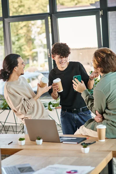 Молодые коллеги улыбаются во время кофе-брейка в светлом офисе, 20-летний мужчина показывает смартфон — стоковое фото