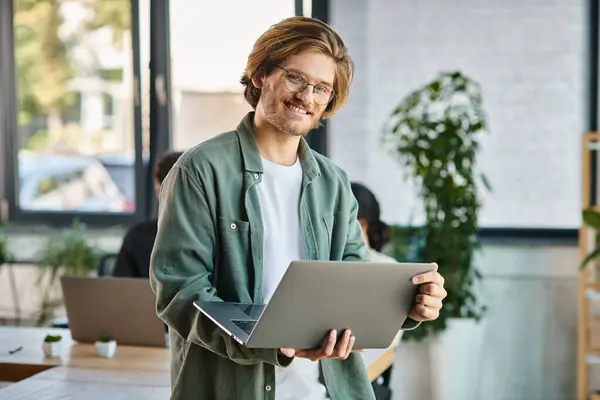 Cheerful young businessman in glasses using laptop and standing in a modern coworking space — Stock Photo