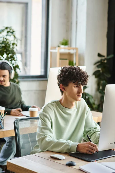 Joven hombre rizado comprometido en el trabajo de retoque en una tableta gráfica con lápiz lápiz óptico cerca de colega - foto de stock