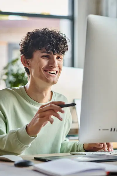 Hombre alegre con el pelo rizado comprometido en el trabajo de retoque con lápiz lápiz lápiz en la mano mirando monitor - foto de stock