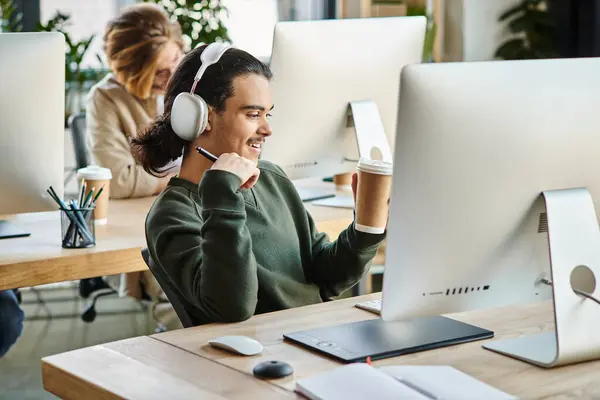 Professionnel masculin joyeux dans les écouteurs tenant café pour aller et travailler sur le projet à son bureau — Photo de stock
