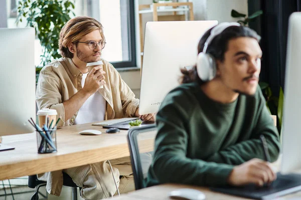 Pensive professional in glasses holding coffee to go and looking at monitor near blurred coworker — Stock Photo