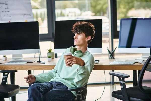 Молодой кудрявый мужчина сидит в офисном кресле и держит кофе, чтобы пойти, пост члена производственной группы — стоковое фото