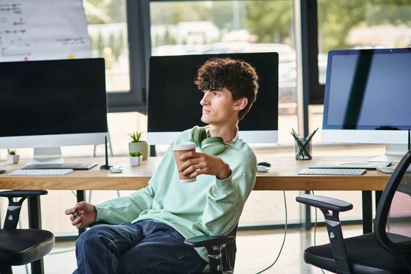 Young curly-haired man sitting in office chair and holding coffee to go, post production team member — Stock Photo