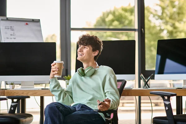 Счастливый молодой человек с наушниками, сидя в офисном кресле и держа кофе, пост производства — стоковое фото