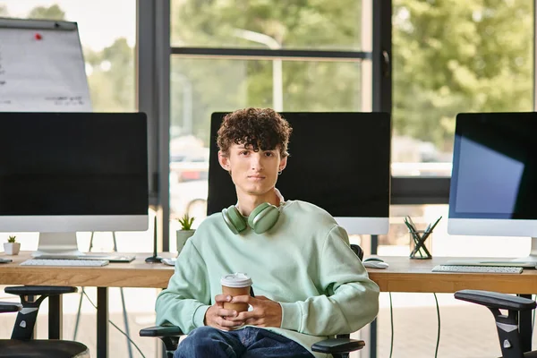 Giovane con i capelli ricci seduto sulla sedia dell'ufficio e tenendo il caffè, membro del team di post produzione — Foto stock