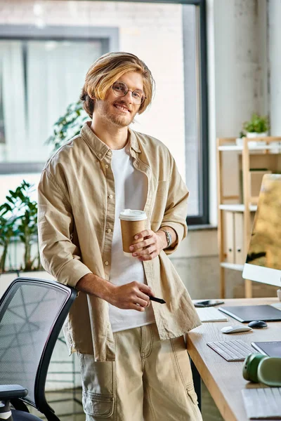 Happy young man in glasses holding coffee and stylus pen in modern office setup, post production — Stock Photo