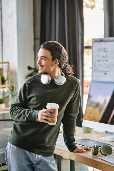 Расслабленный молодой человек, стоящий со стилусом ручкой и кофе и улыбающийся в пост-продакшн рабочем пространстве — стоковое фото