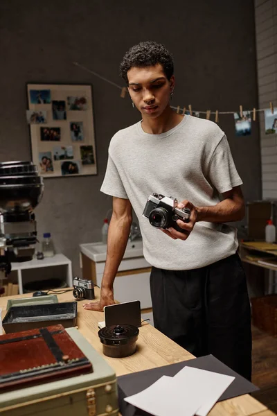 Jeune homme noir Examiner intensément un appareil photo analogique tout en se tenant dans un laboratoire photo, photographie de film — Photo de stock
