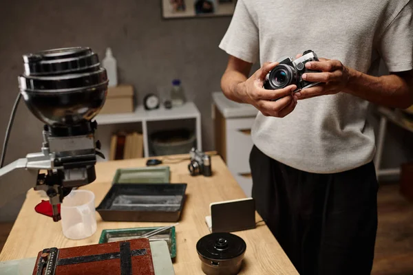 Hombre negro recortado Sujetando intencionalmente una cámara analógica mientras está de pie en un laboratorio de fotografía, fotografía de película - foto de stock