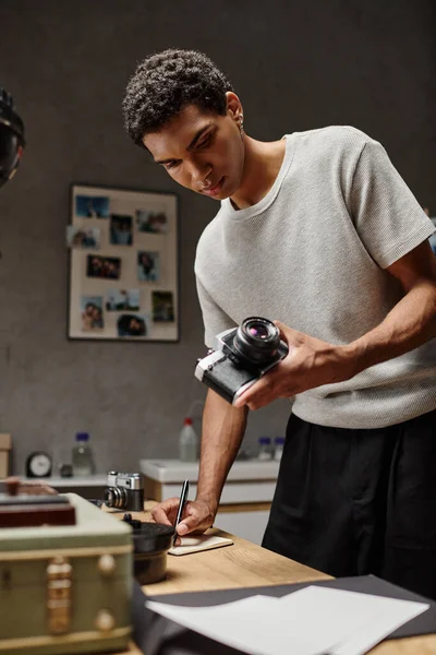 African American photographer immersed in writing holding his analog camera in photo studio — Stock Photo