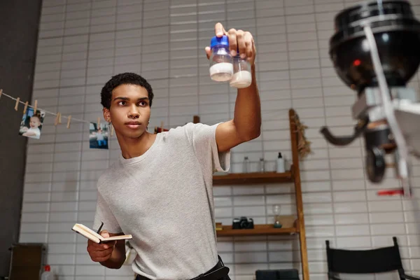 Bonito fotógrafo afro-americano segurando garrafas com pó de desenvolvimento de filme e notebook — Fotografia de Stock