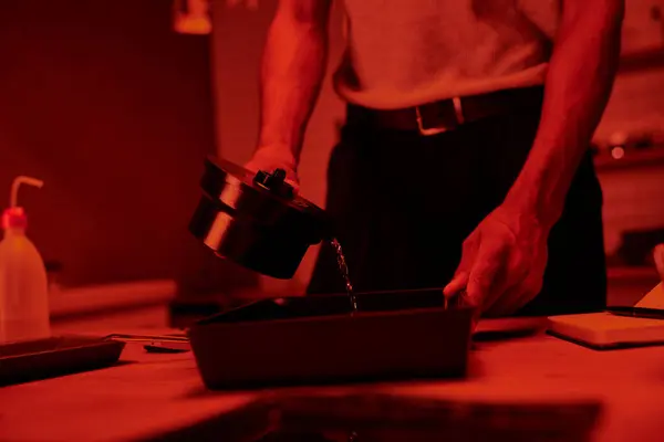 Ausgeschnittener Fotograf gießt Entwickler in ein Tablett zur Filmbearbeitung in Dunkelkammer mit Rotlicht — Stockfoto