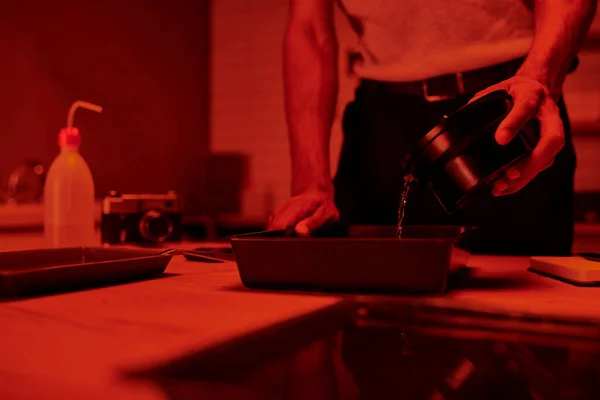 Ausgeschnittene Ansicht des Fotografen gießt Entwickler in Tablett zur Filmbearbeitung in roter Dunkelkammer — Stockfoto