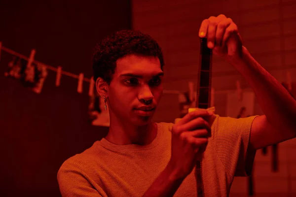 Afro-américain photographe examine développé bande de film, baigné dans la chambre noire avec la lumière rouge — Photo de stock