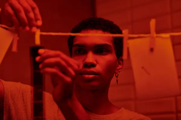 Photographe afro-américain accroche bande de film fraîchement développé dans une chambre noire rouge-éclairé, intemporel — Photo de stock