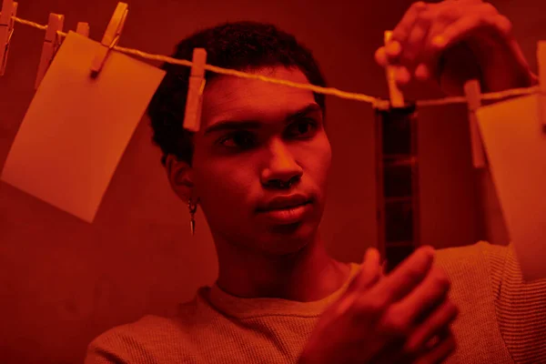 Молодий афроамериканський чоловік вішає свіжо розвинену плівкову стрічку в червоно-світлій темній кімнаті, безстроково — стокове фото