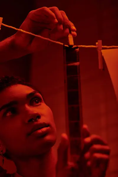 Jeune homme afro-américain accroche bande de film fraîchement développé dans une chambre noire rouge-éclairé, intemporel — Photo de stock