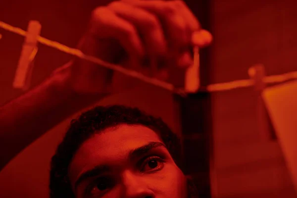 Jovem homem americano africano pendura tira de filme recém-desenvolvido em uma câmara escura vermelho-iluminada, nostalgia — Fotografia de Stock