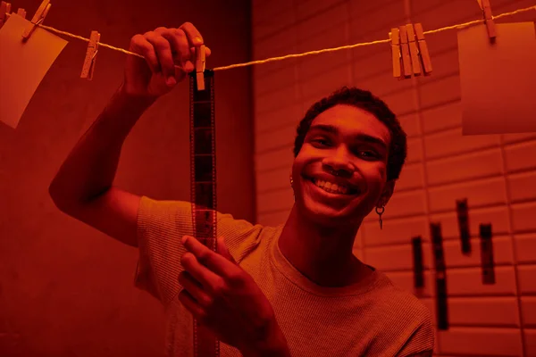 Cheerful african american man hanging freshly developed film strip  in a red-lit darkroom, nostalgia — Stock Photo